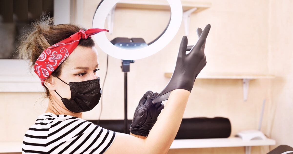 Female tattoo artist with ring light putting on rubber gloves with mask on.