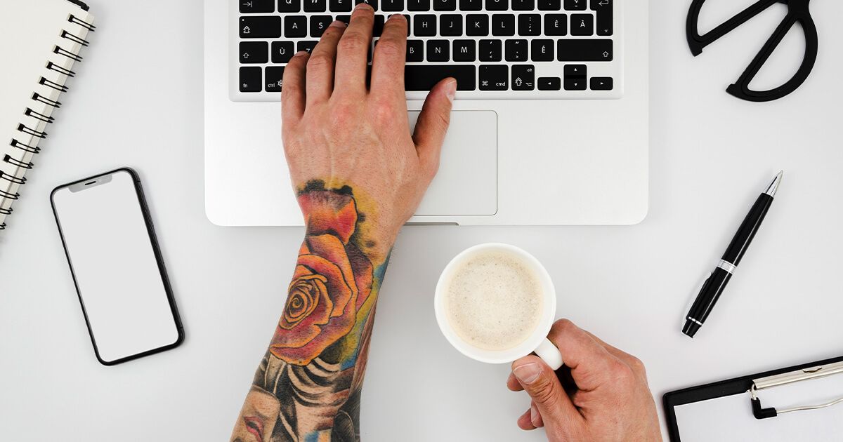 Man typing on laptop keyboard with coffee. Arm with tattoo sleeve.